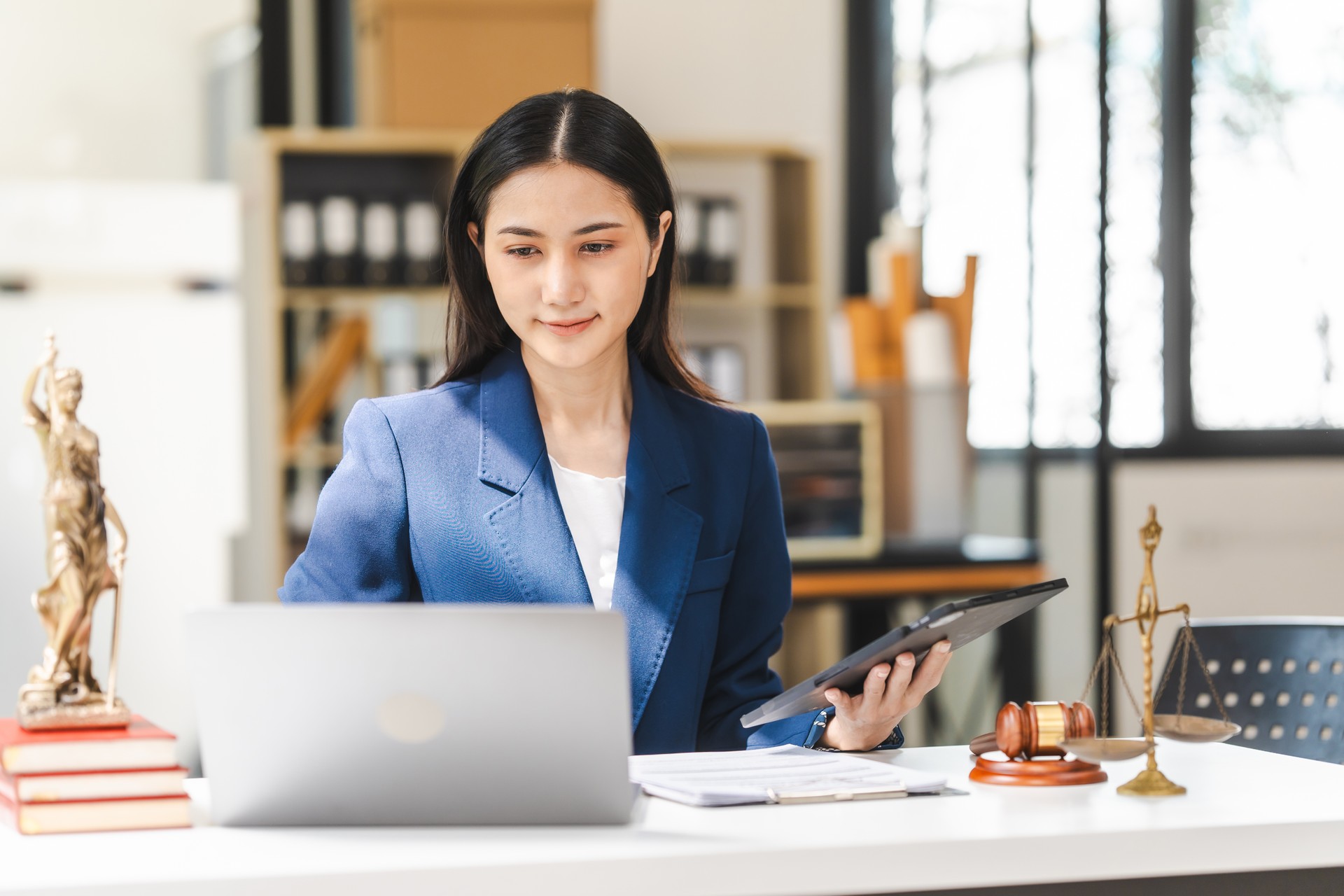 Young attractive Asian female lawyer in formal suit works on tablet with laptop, legal books